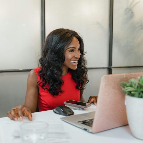 Professional black woman smiling at desk using laptop and smartphone in office.